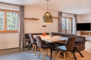 Een moderne eetruimte met een houten tafel, donkere gestoffeerde stoelen, een hangende hanglamp en een televisie op de achtergrond.