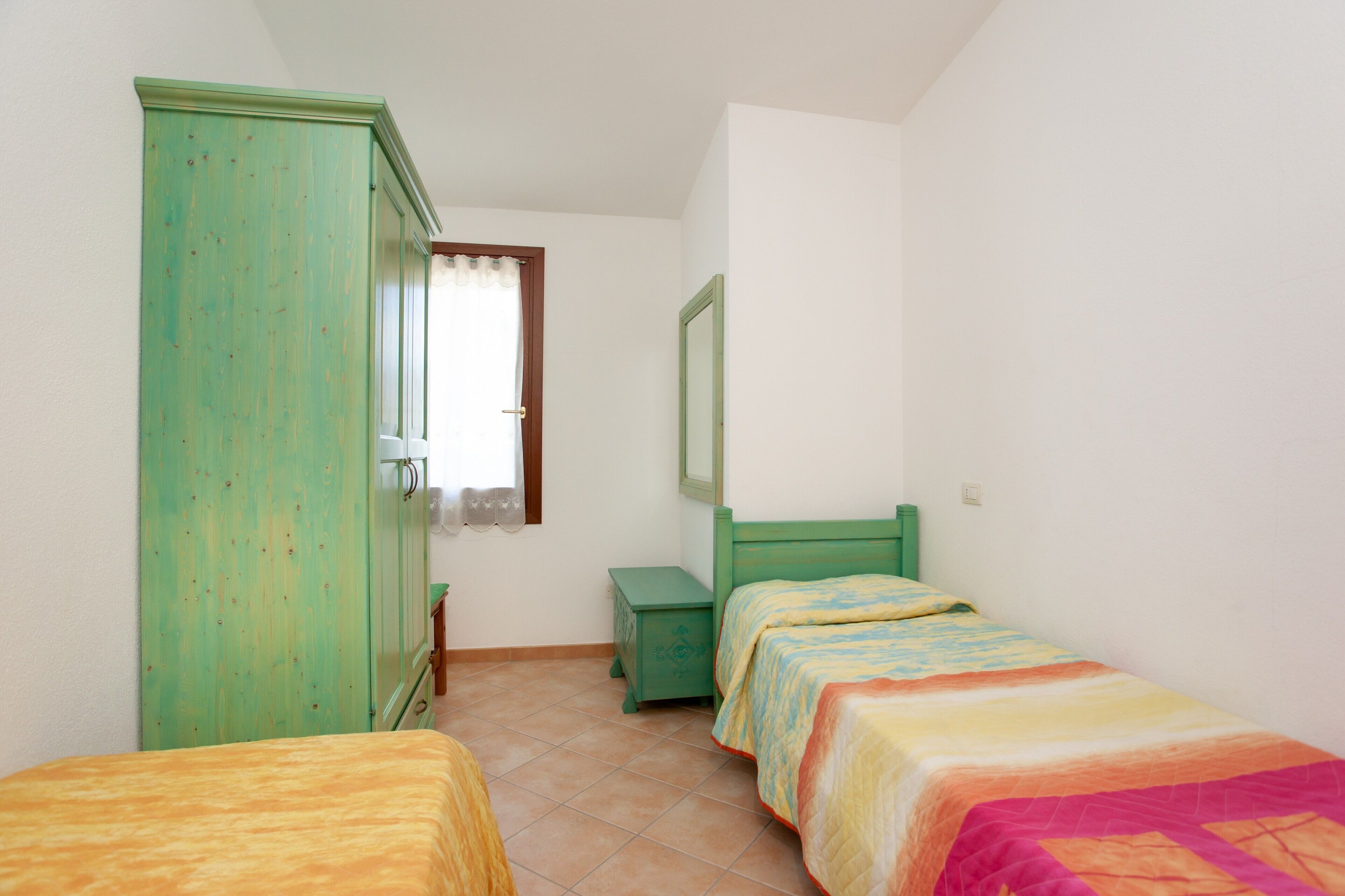 A bedroom with two single beds, a green wardrobe, a small bedside table, and a window with a curtain.