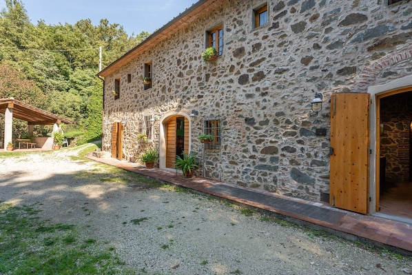 Un bâtiment en pierre avec une porte en bois, un espace extérieur abrité avec des sièges et des plantes en pot.