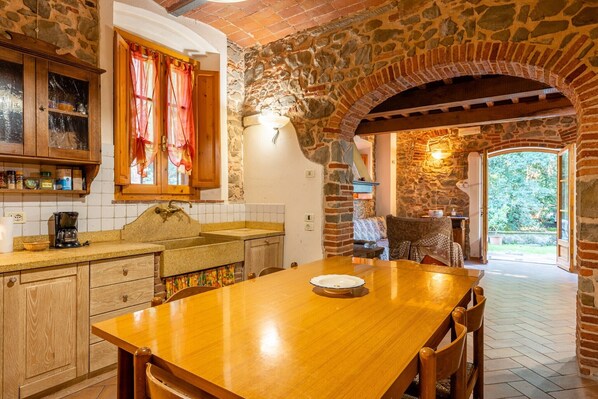A stone-walled kitchen with a wooden table and chairs, a window with curtains, and a view of the outdoors.