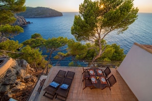 A terrace with a table and chairs, overlooking a coastal area with trees and the sea.