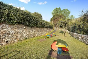 Un mur de pierres, une zone herbeuse avec des jeux pour enfants et des arbres en arrière-plan.