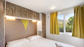 A neatly made bed with white linens, a wooden headboard, a bedside table with a lamp, and a window with curtains. - 'Mobile Home Alloro 165' with Pool, Private Terrace & Wi-Fi (Pietra Ligure)