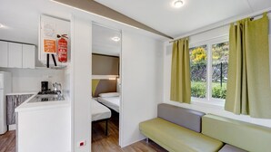 A compact room with a kitchenette, a bed, a bench, and a window with curtains. - 'Mobile Home Alloro 165' with Pool, Private Terrace & Wi-Fi (Pietra Ligure)