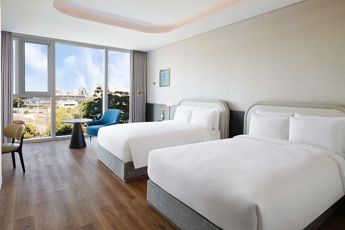 A hotel room with two beds, a chair, a small table, and a view of the city through large windows.