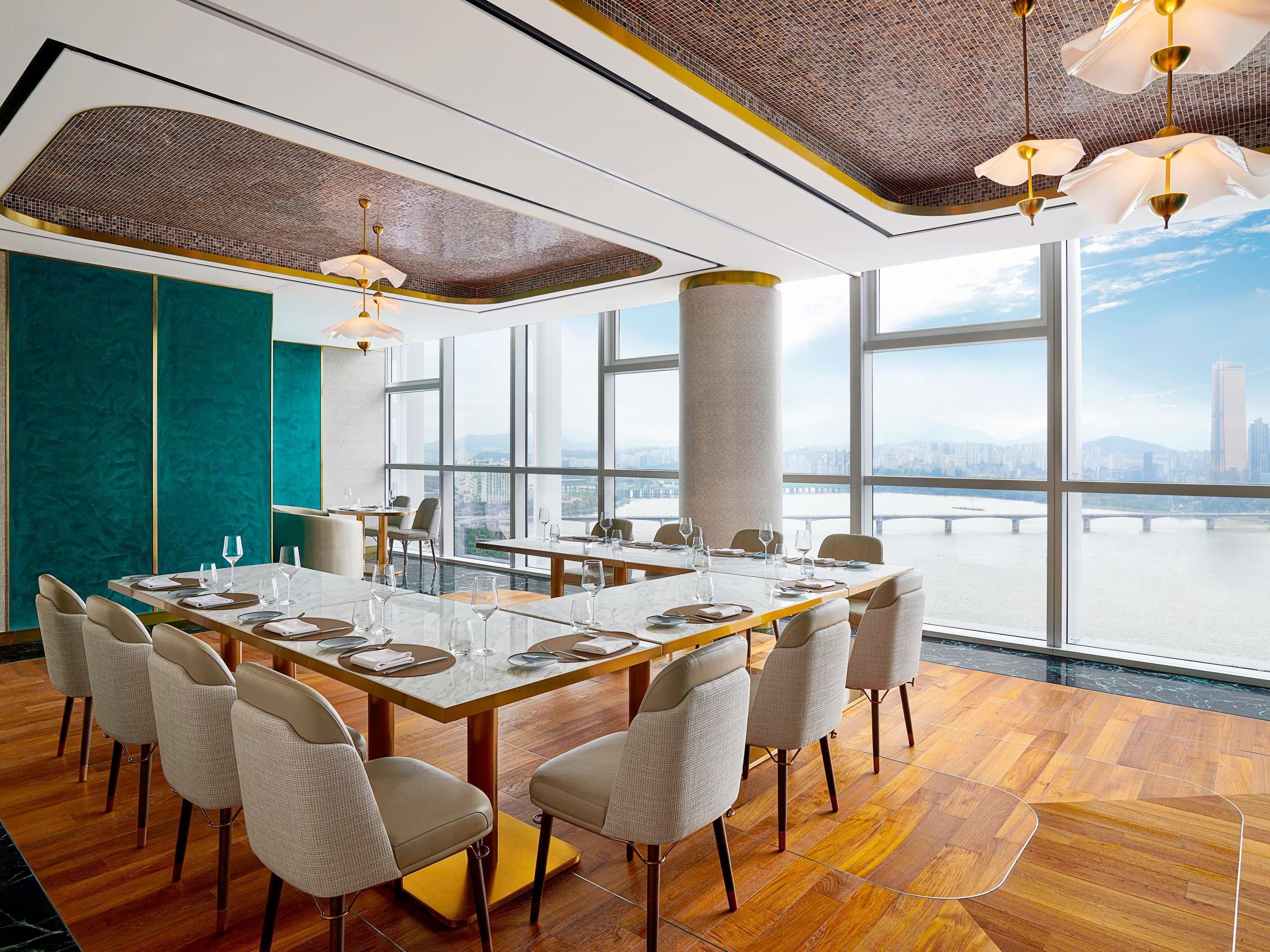A modern dining area with a large table, chairs, and large windows offering a city view.
