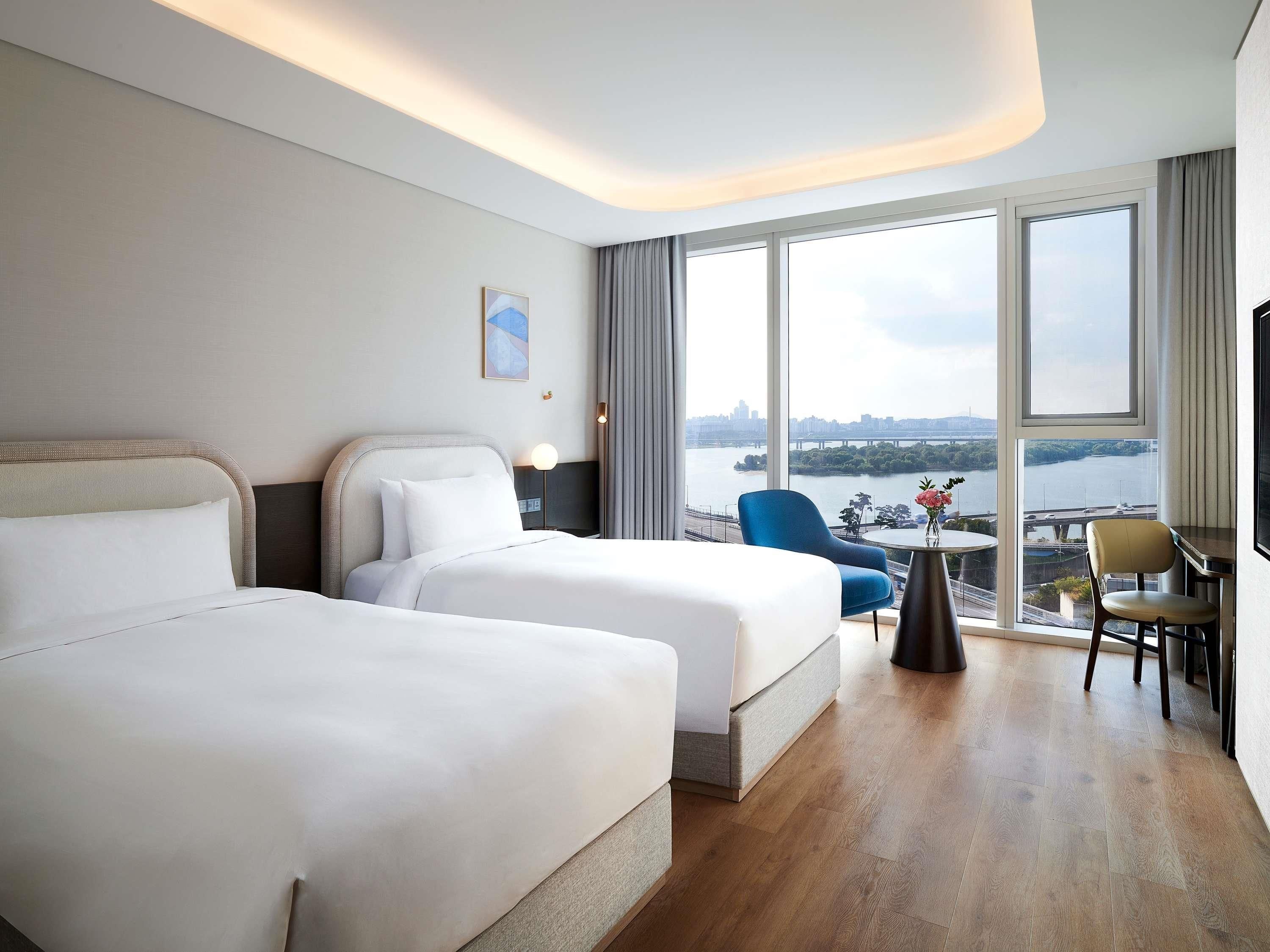 A hotel room with two beds, a view of a river and cityscape, and a small table with a vase.