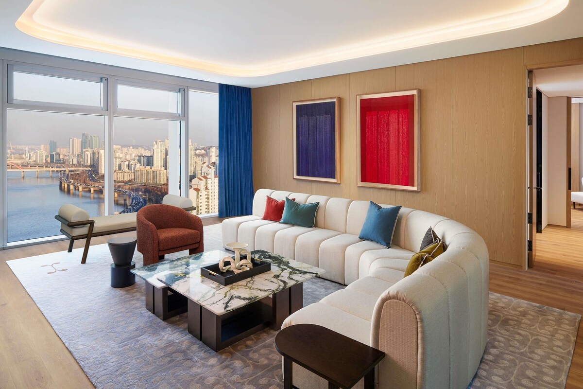 A modern living room with a large sofa, a coffee table, and a view of a cityscape through large windows.