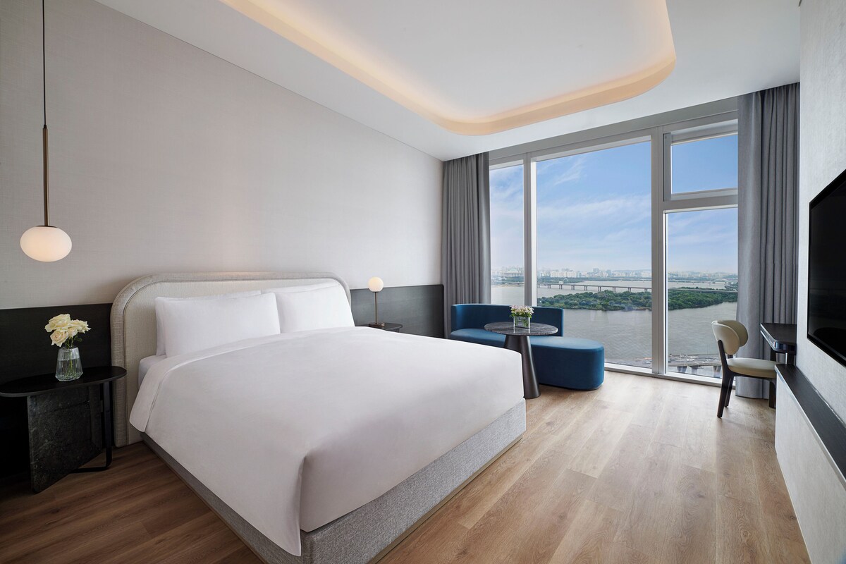 A modern hotel room with a large bed, a desk with a lamp, a small table with flowers, a blue chair, and a TV.