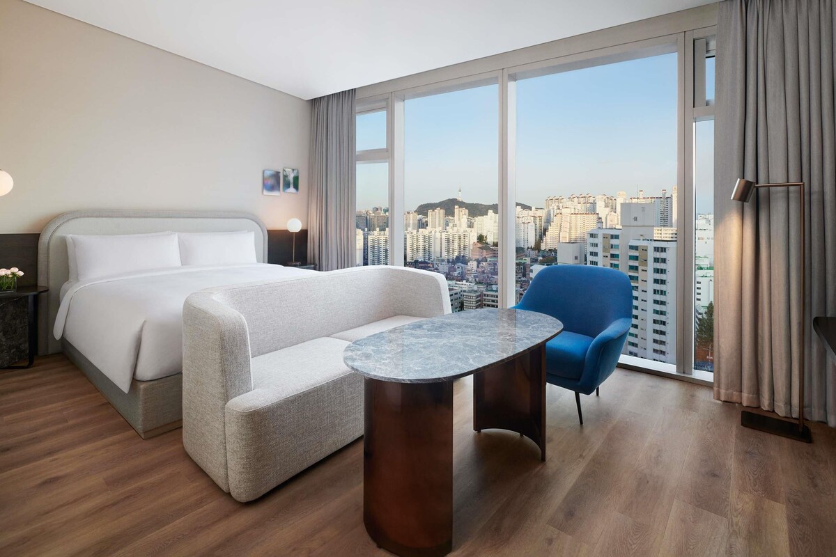 A hotel room with a bed, a seating area with a table, a chair, and a view of the cityscape through large windows.