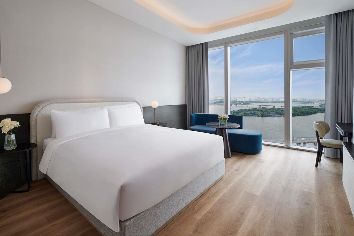 A modern hotel room with a large bed, a desk, a chair, and a view of a river and cityscape.