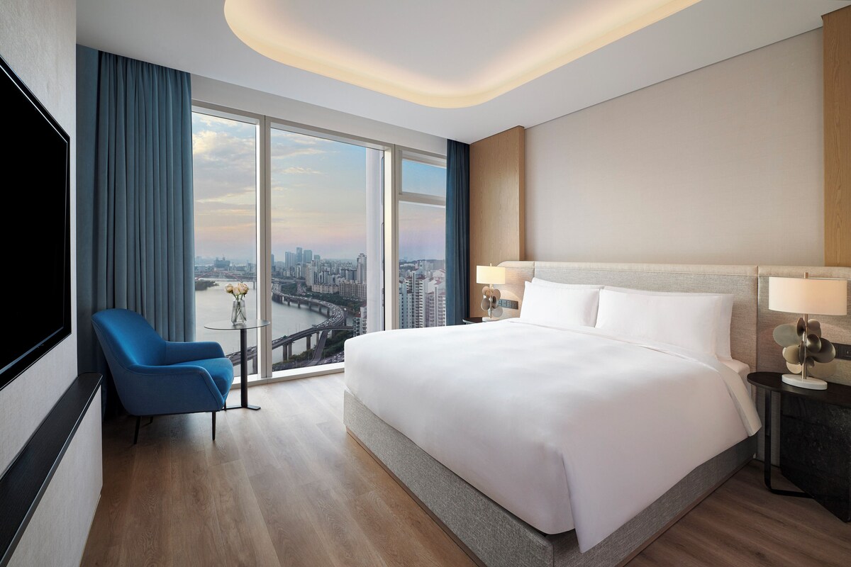 A modern hotel room with a large bed, a blue armchair, a small table with a vase, and a city view through large windows.