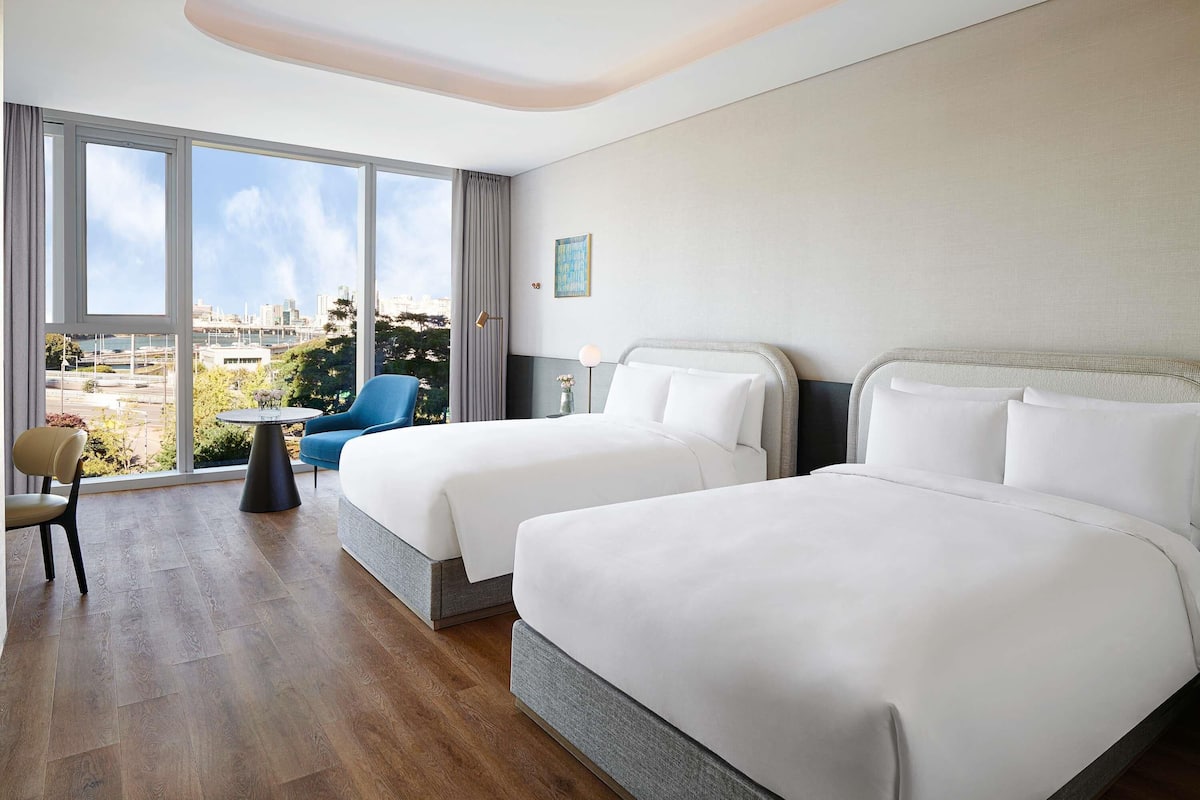 A hotel room with two beds, a chair, a small table, and a view of the city through large windows.