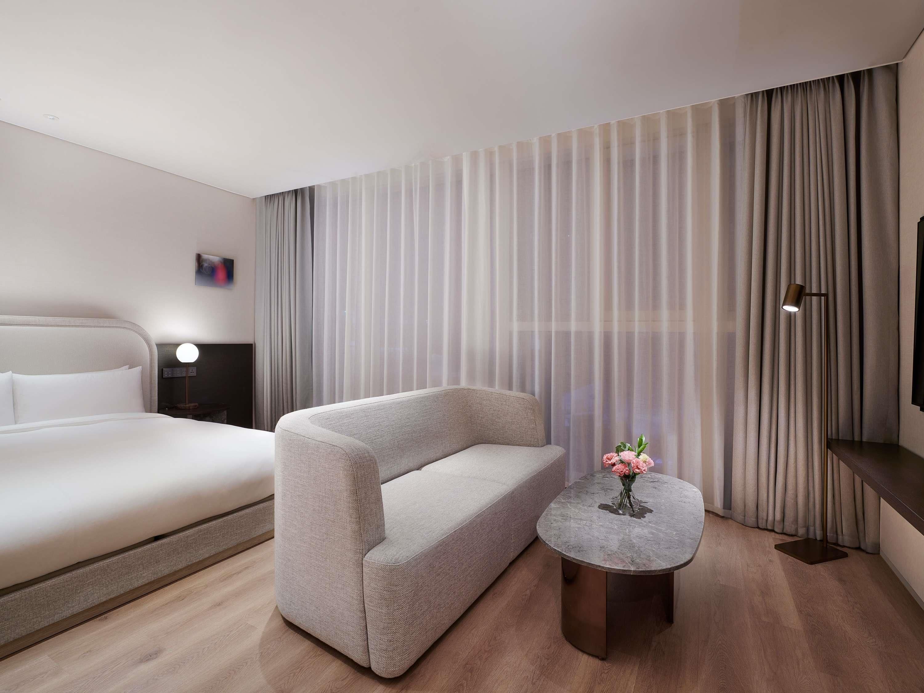 A modern hotel room with a bed, a sofa, a small round table with a vase of flowers, and a TV on a stand.
