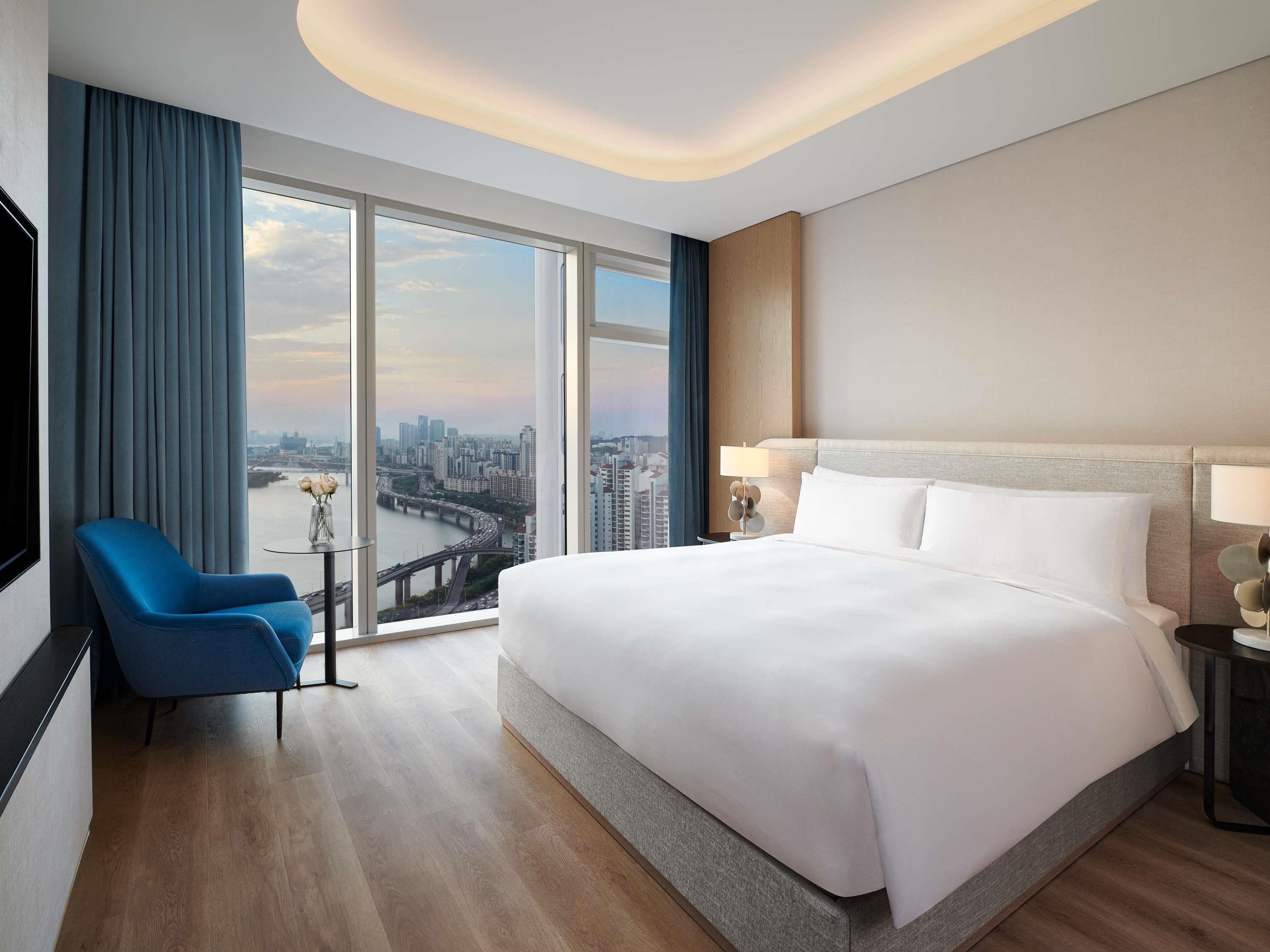 A hotel room with a large bed, a blue armchair, a small table with a vase, and a view of a cityscape through large windows.