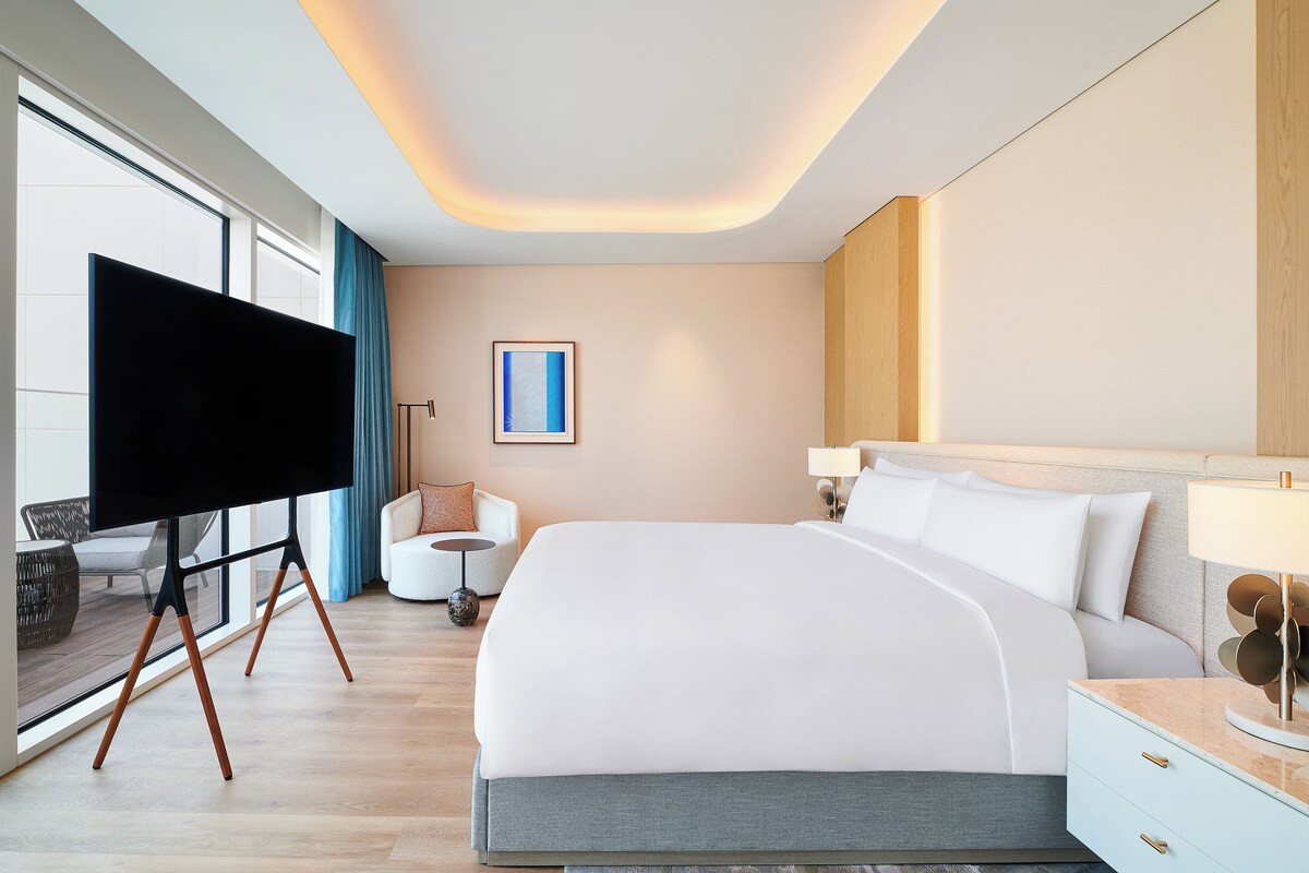 A modern hotel room with a large bed, a flat-screen TV, a small round table, and a view of the outdoors.