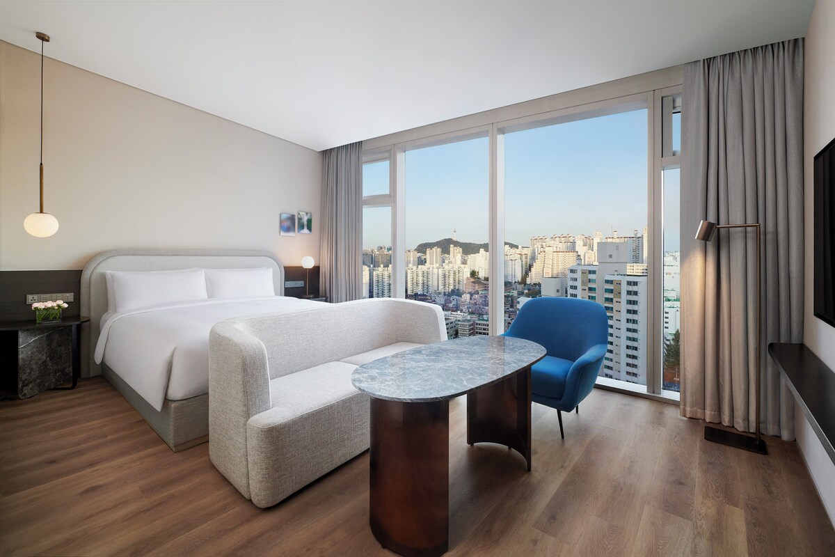 A modern hotel room with a large bed, a seating area with a round table, and a city view through large windows.