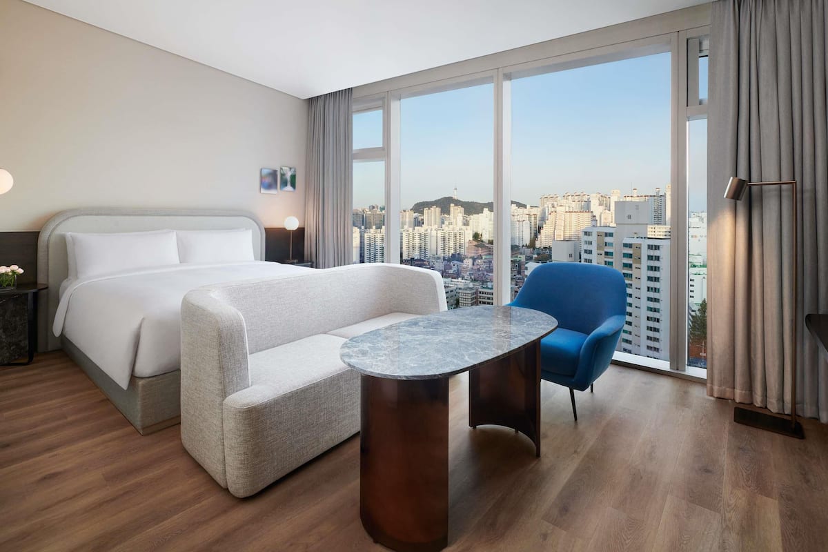 A hotel room with a bed, a seating area with a table, a chair, and a view of the cityscape through large windows.