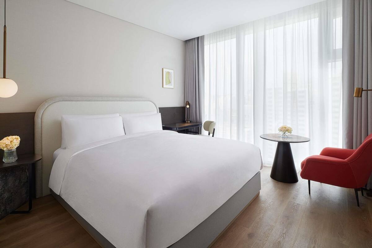 A hotel room with a large bed, a desk, a chair, and a view of the city through sheer curtains.