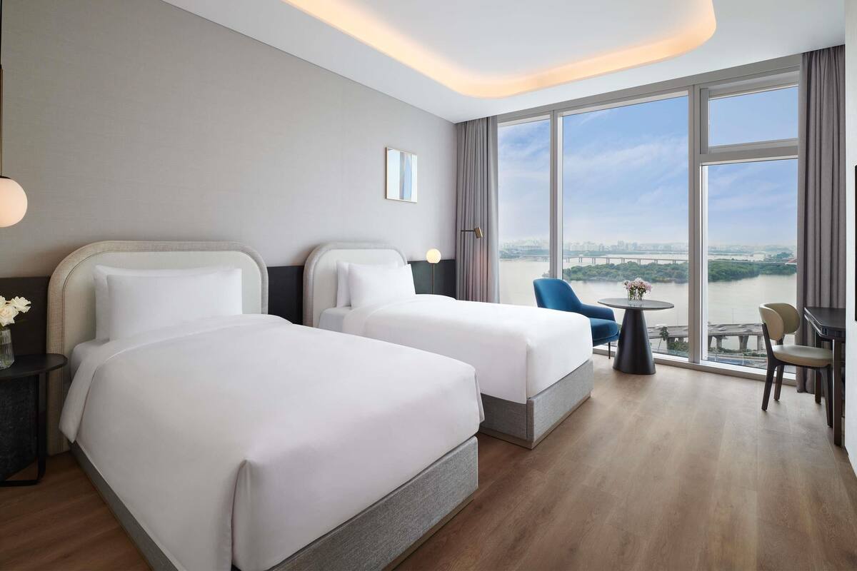 A hotel room with two beds, a view of a river, a dining area with a table and chairs, and a large window.