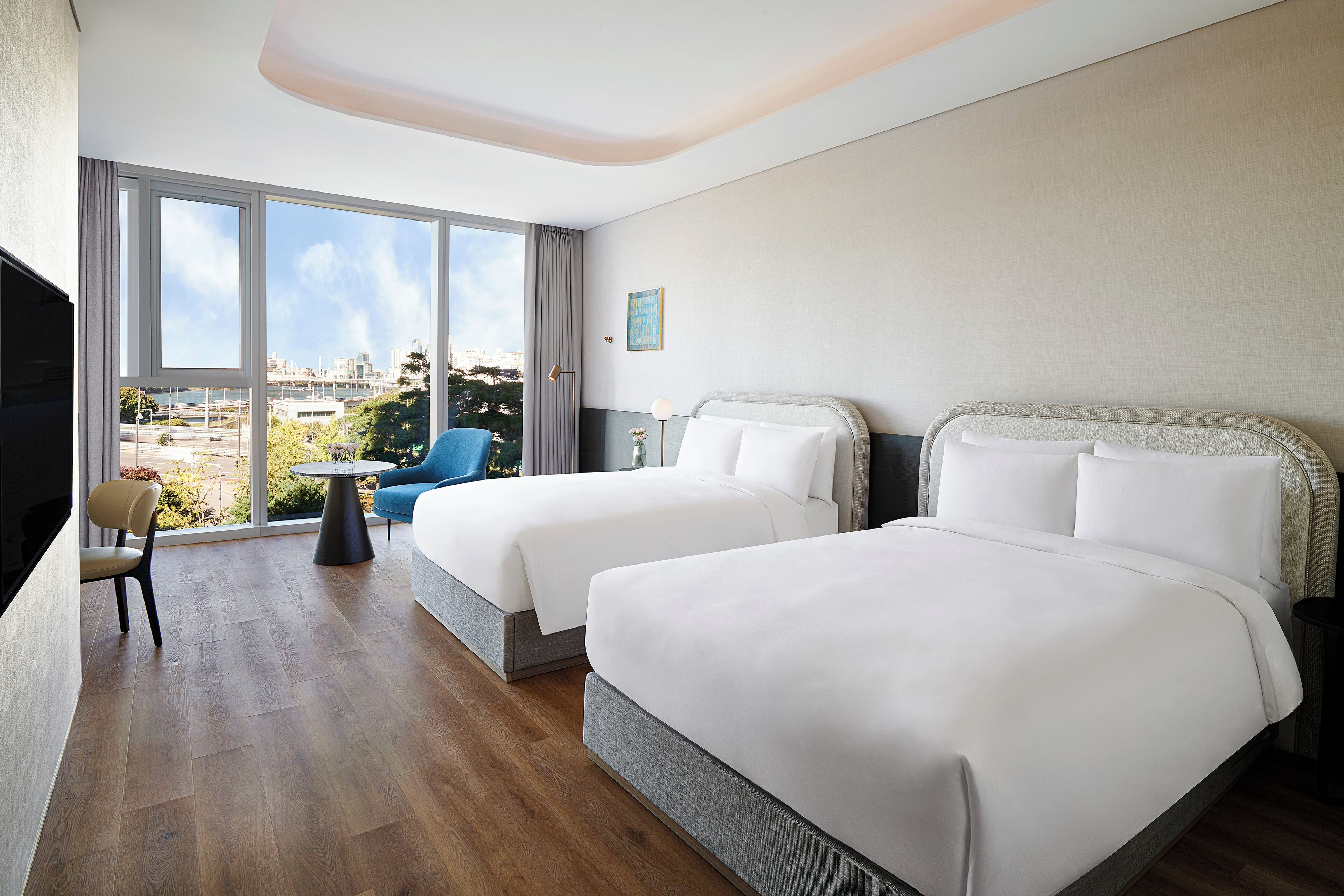 A hotel room with two beds, a large window, a chair, and a small table.