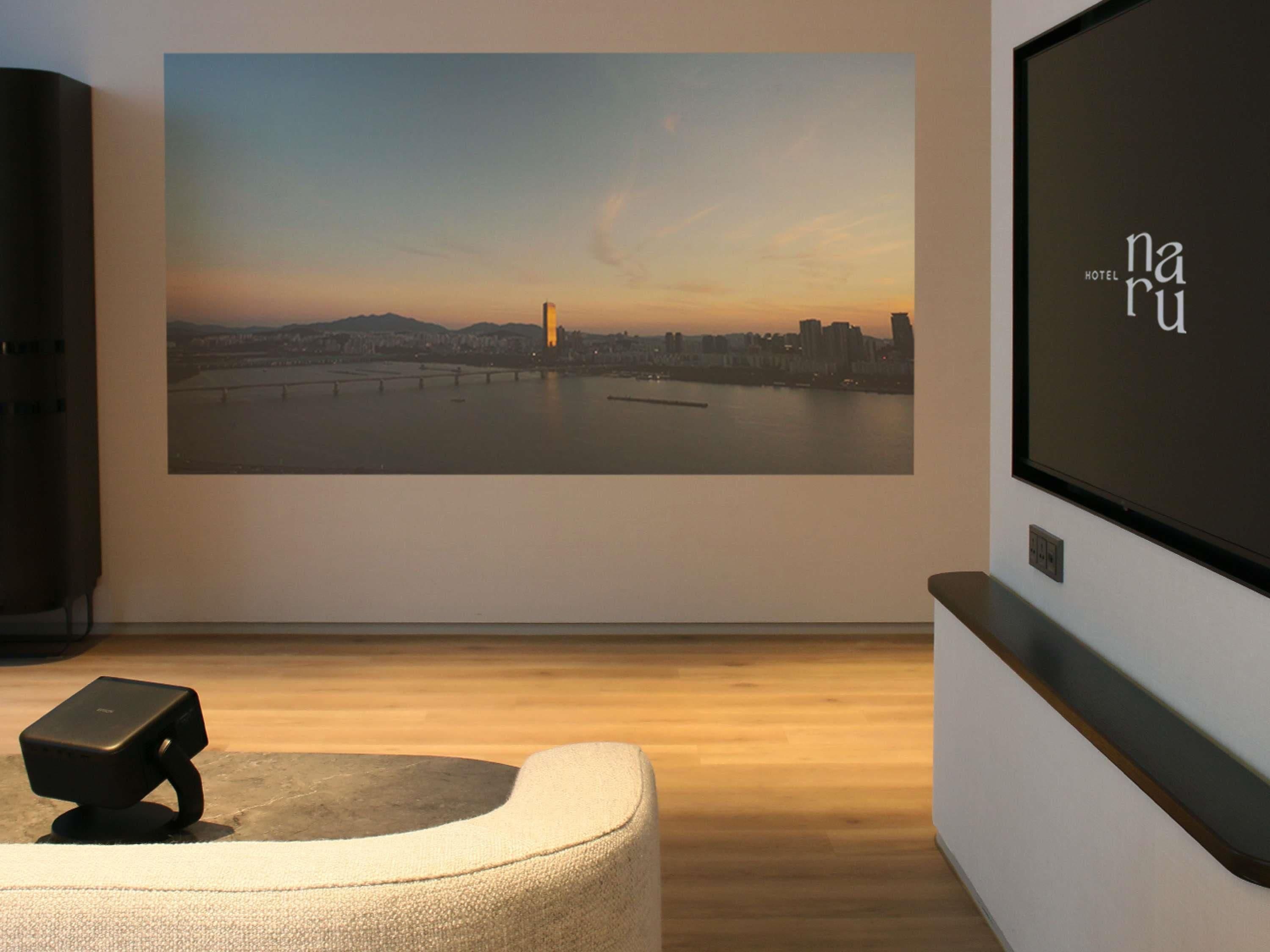 Ein Hotelzimmer mit großem, an der Wand montierten Fernseher, der eine Stadtansicht zeigt, einem Flachbildfernseher an der Wand und einer modernen Sitzecke.
