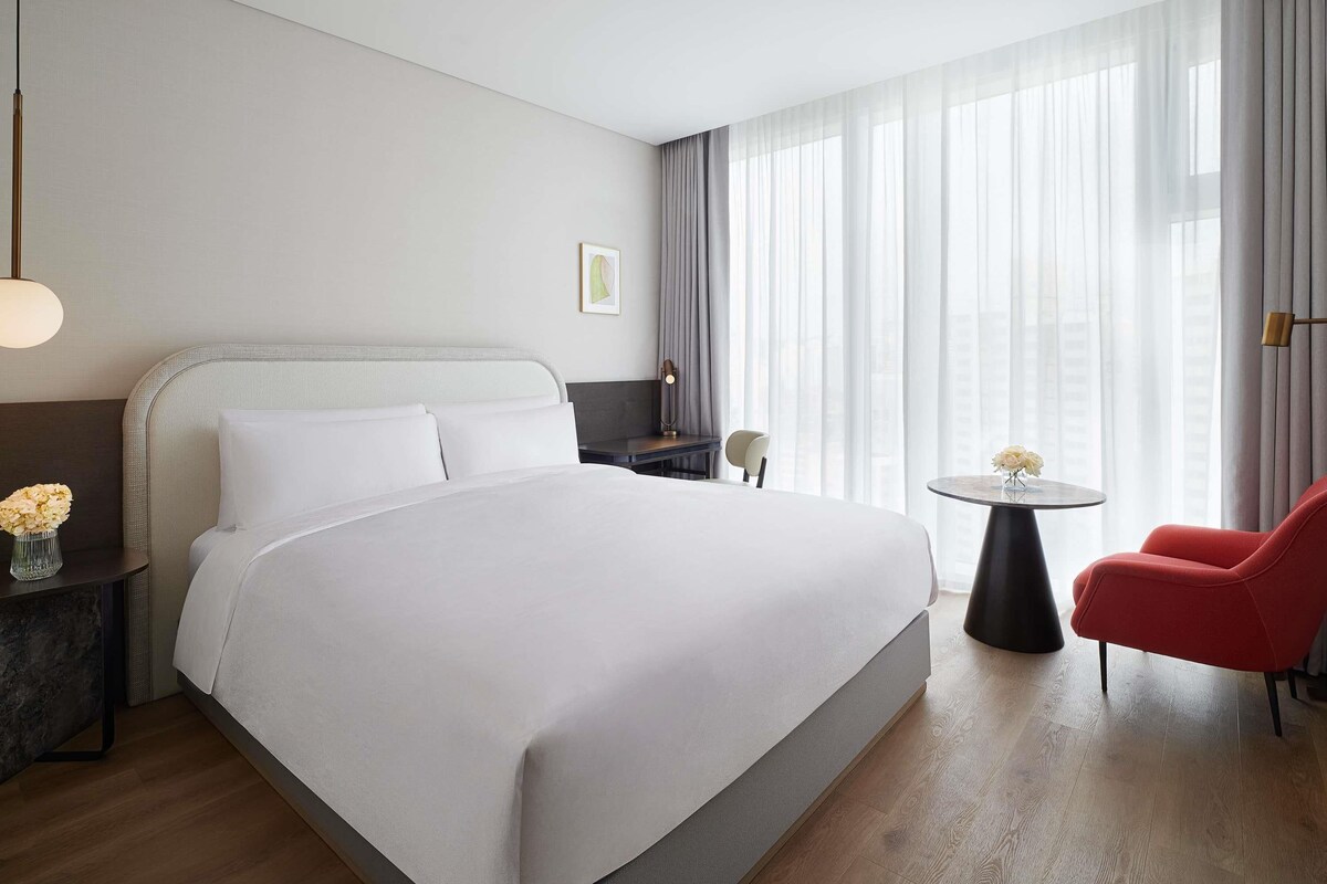 A hotel room with a large bed, a desk, a chair, and a view of the city through sheer curtains.