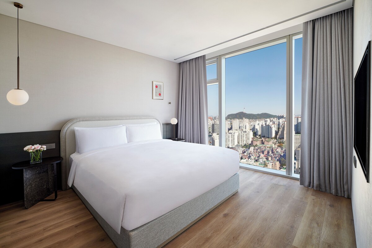 A modern hotel room with a large bed, a nightstand, a vase of flowers, a city view through large windows, and a flat-screen TV.