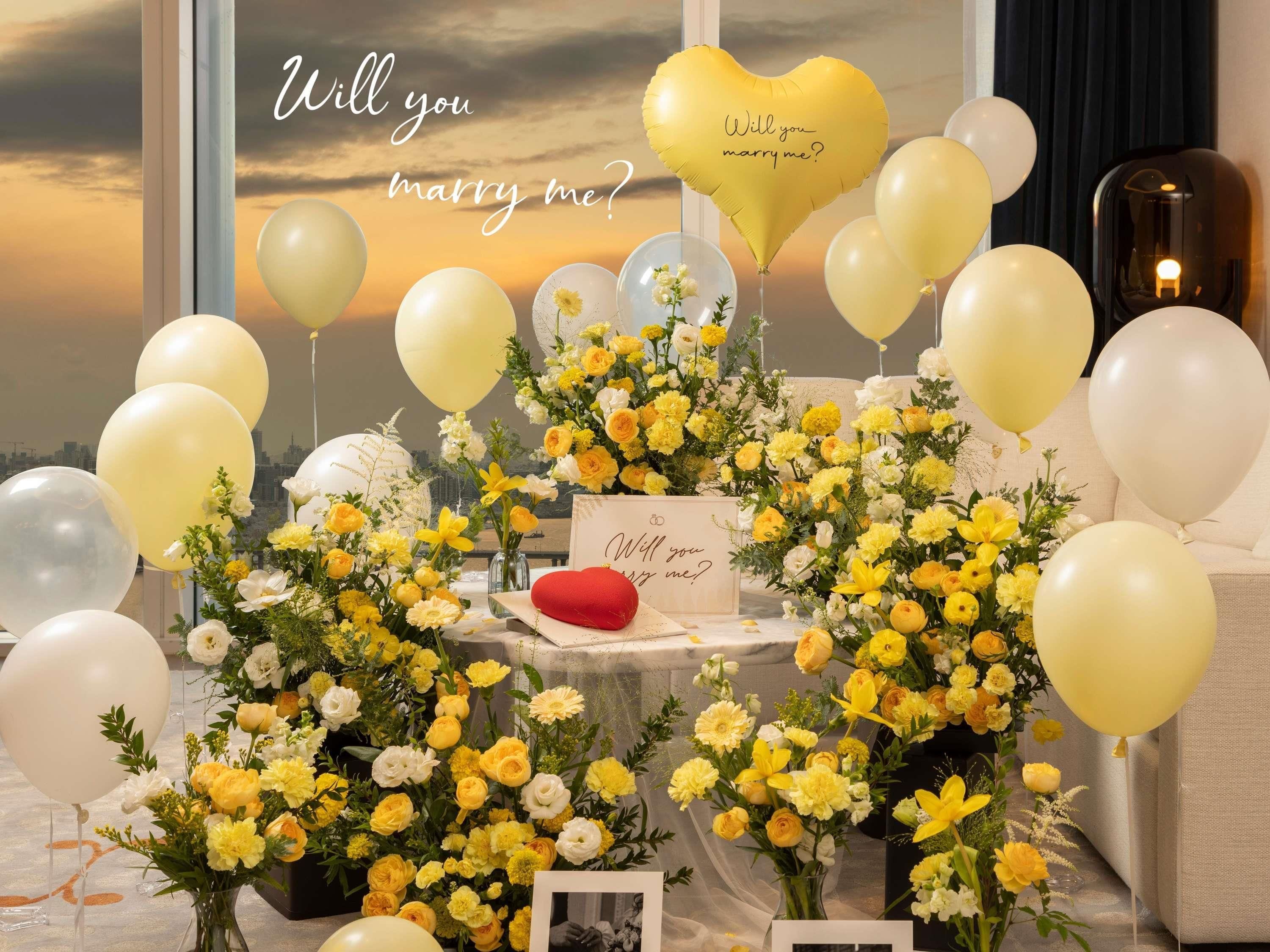 A table with yellow and white flowers, a heart-shaped balloon with 'Will you marry me?' text, and a red cushion.