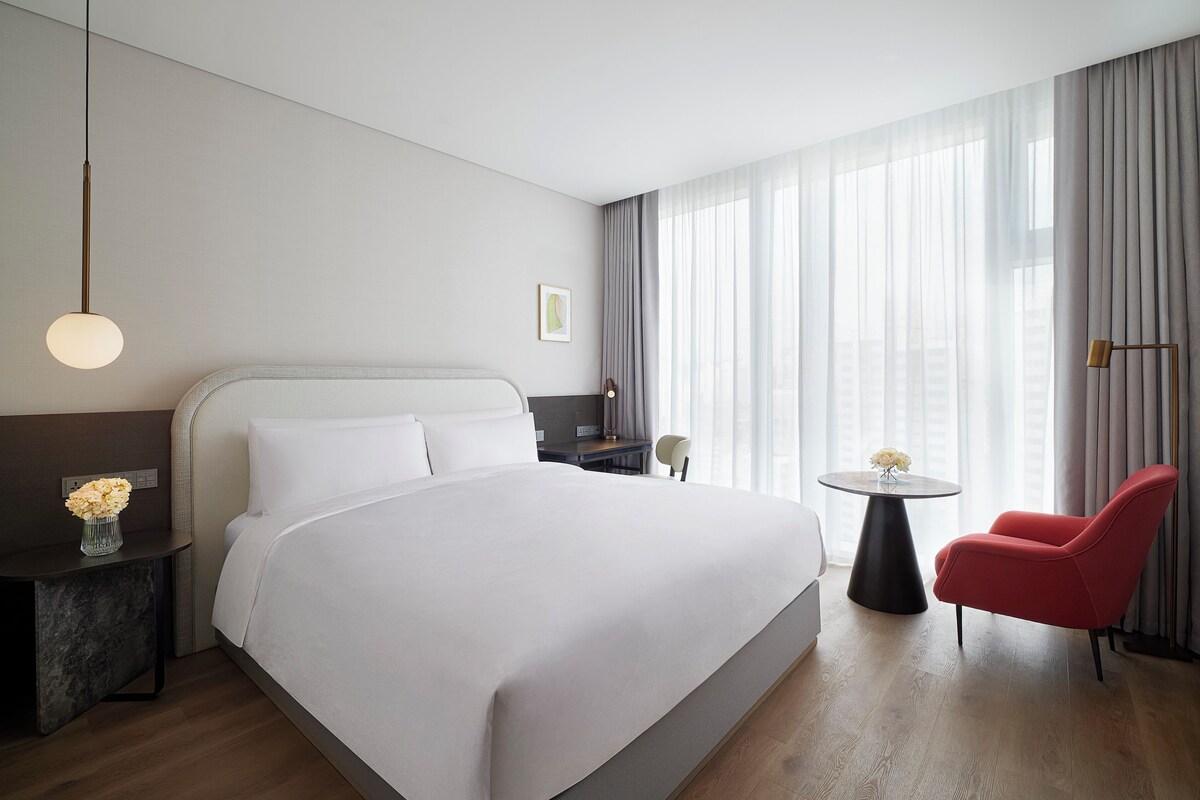 A modern hotel room with a large bed, a small round table with a vase, a red chair, and a view of the city through sheer curtains.
