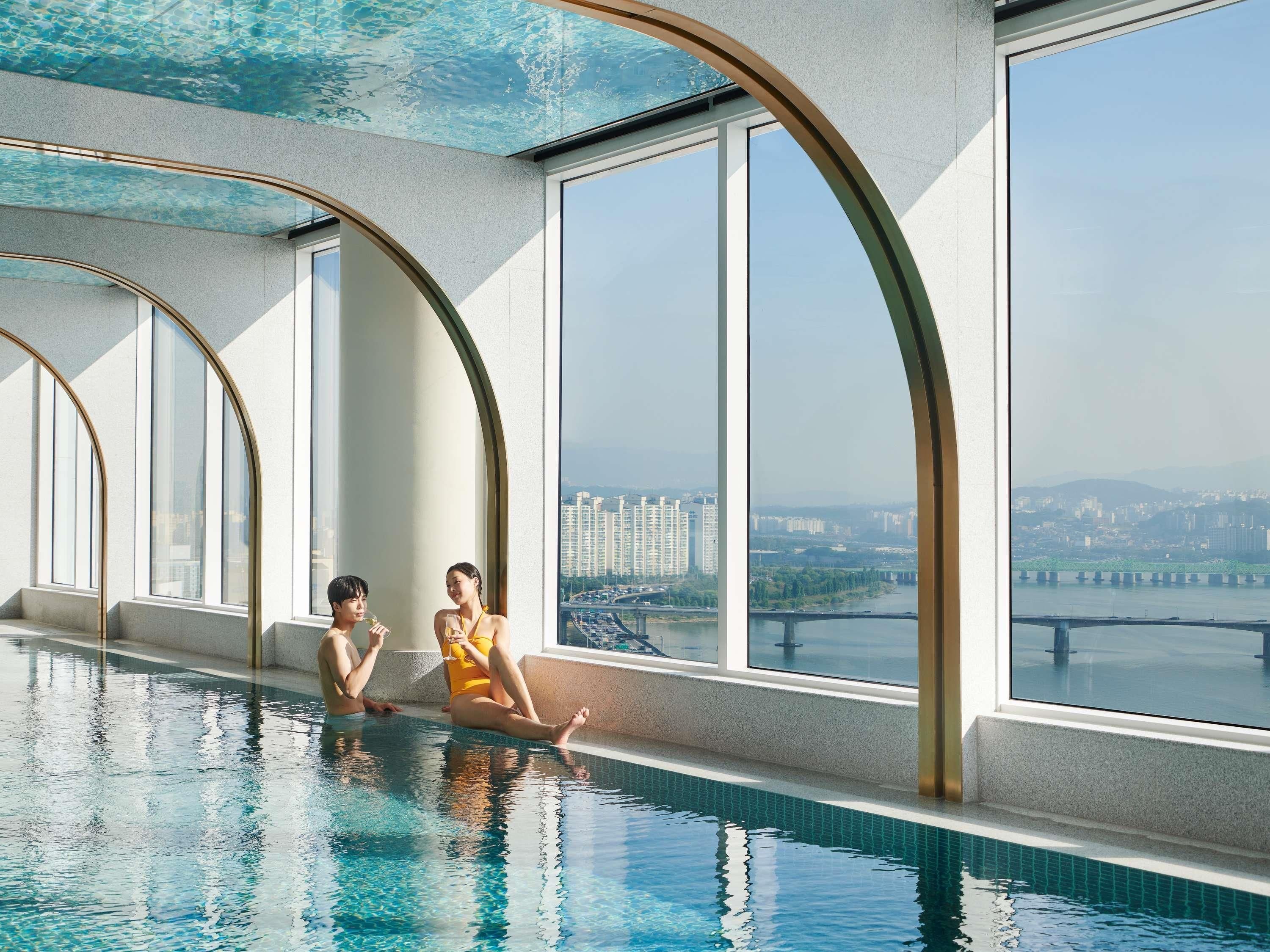 A modern indoor swimming pool with large windows offering views of a cityscape and a bridge.