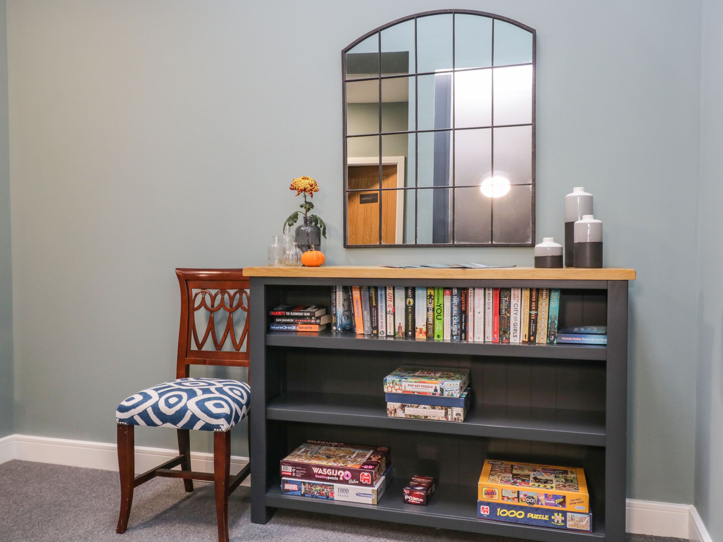 A bookshelf with various books and puzzles, a chair with a patterned cushion, and a mirror on the wall.