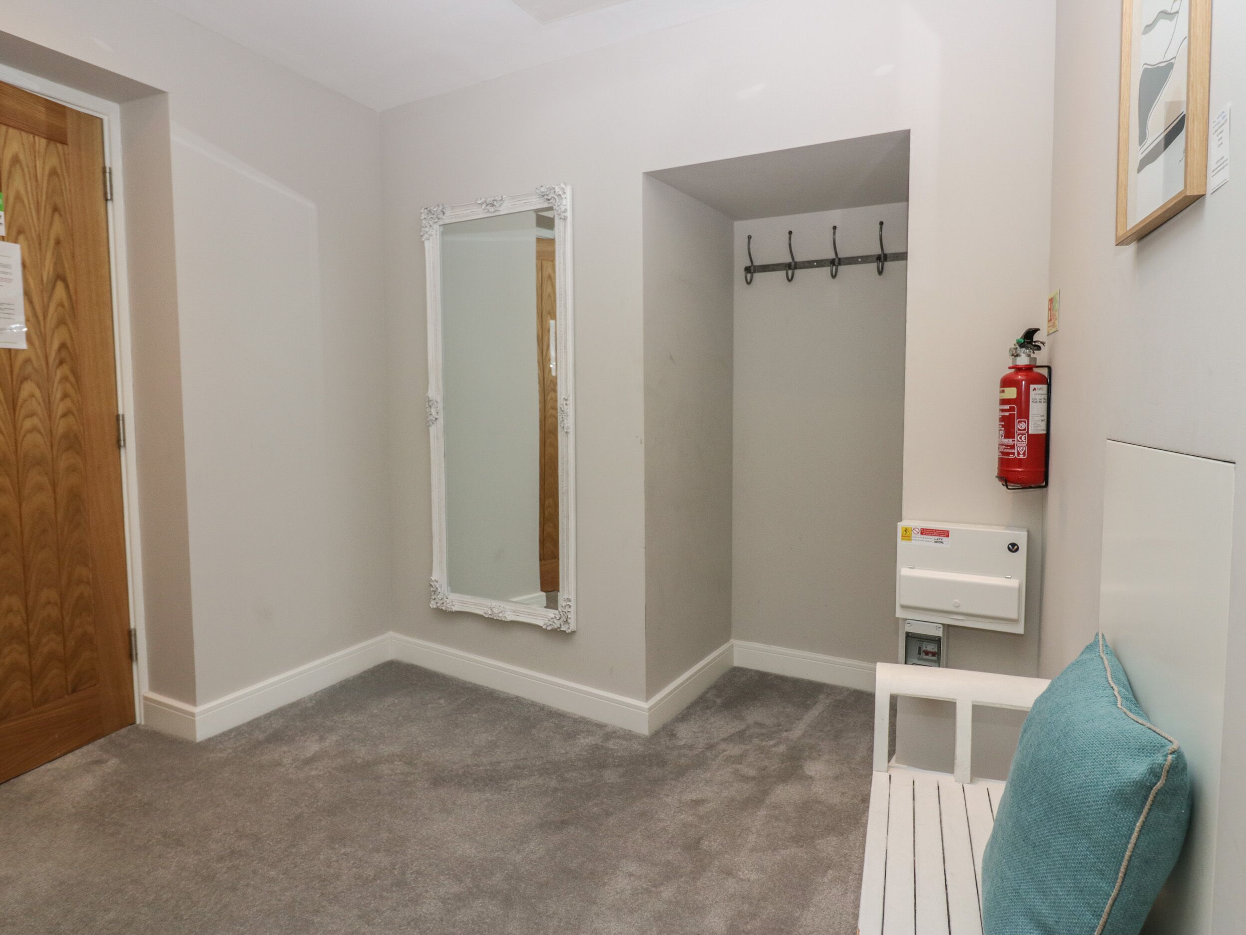 Una habitación con puerta de madera, espejo de marco blanco, banco blanco, cojín color turquesa, extintor y termostato.