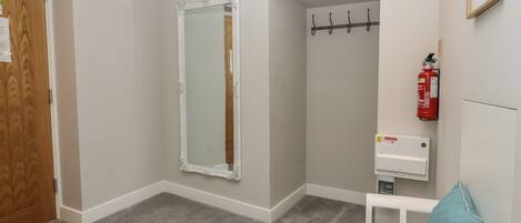 Una habitación con puerta de madera, espejo de marco blanco, banco blanco, cojín color turquesa, extintor y termostato.