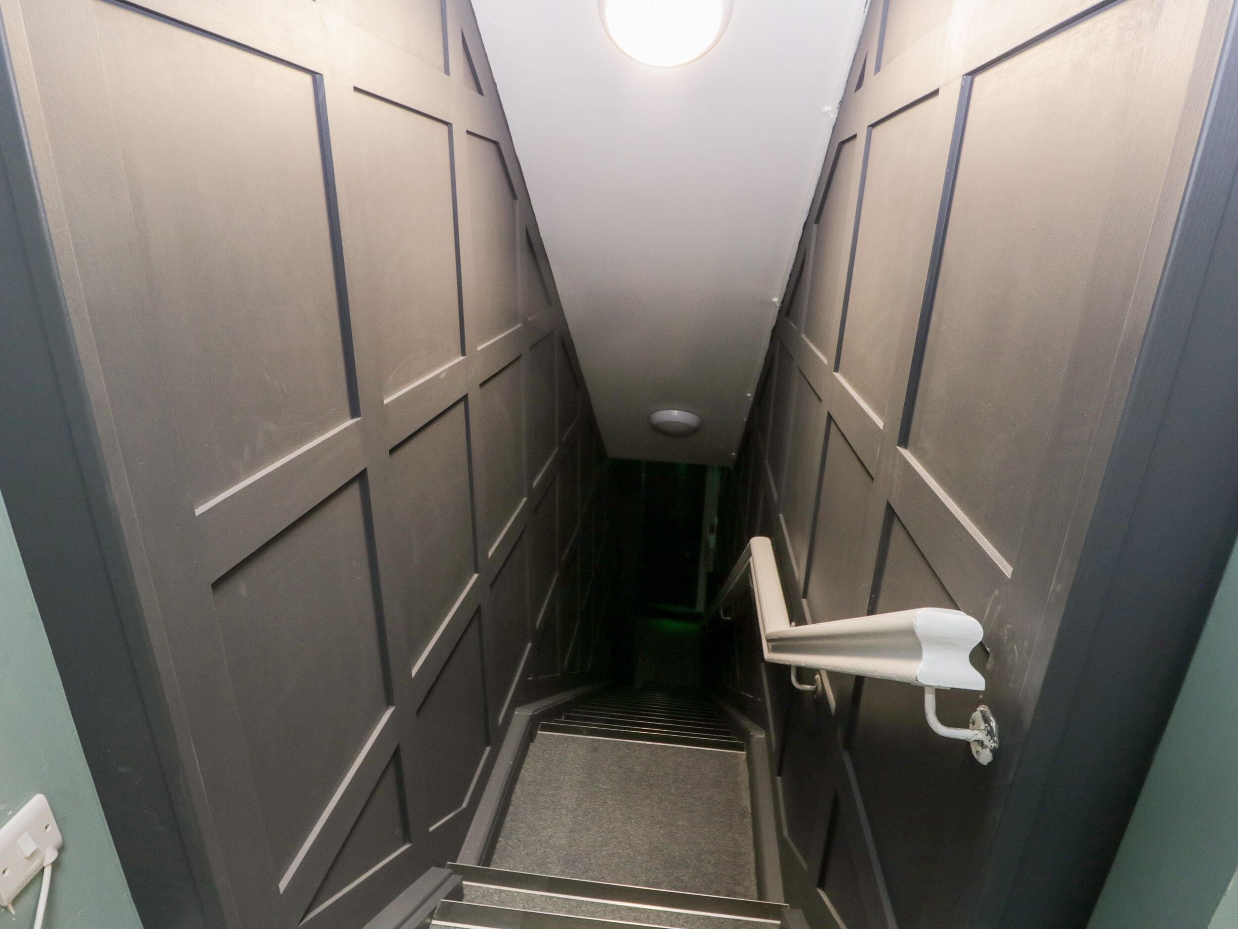 Eine schmale, schwach beleuchtete Treppe mit Handlauf und einer Leuchte am oberen Ende.