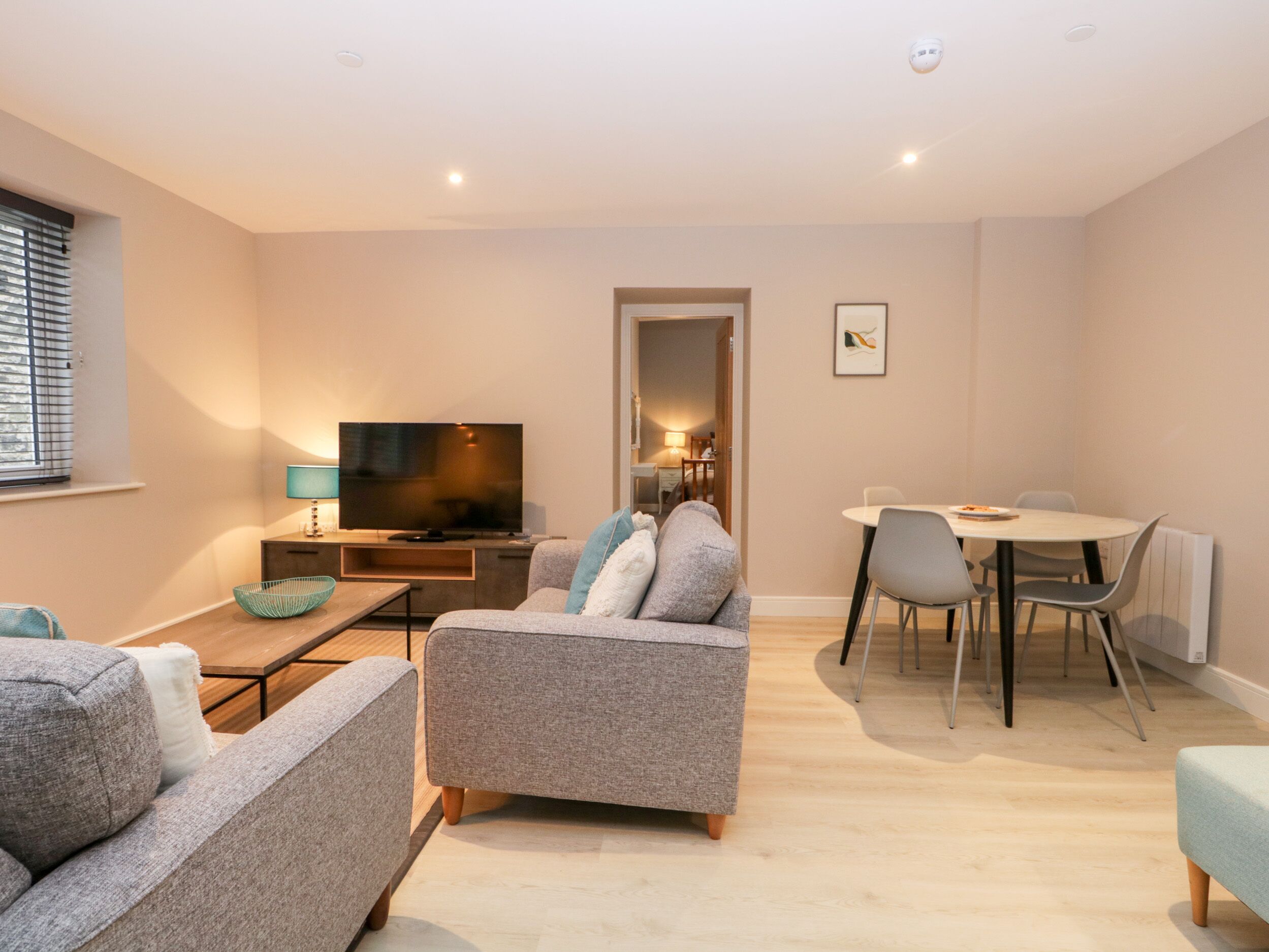 A modern living room with a flat-screen TV, grey sofas, a dining table with chairs, and a small kitchen area.