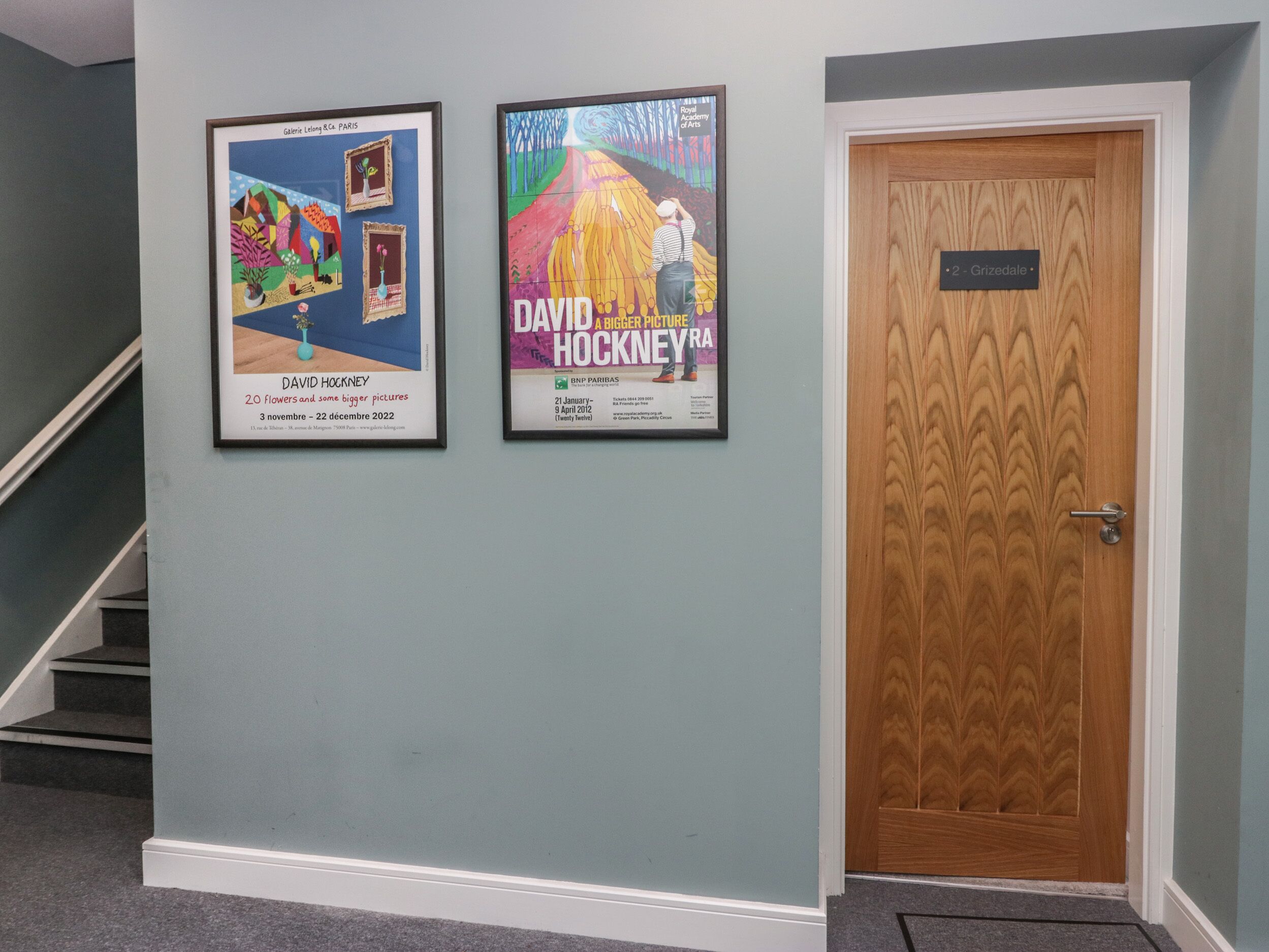 A hallway with a staircase, a wooden door labeled "2 - Grizedale," and two framed posters on the wall.