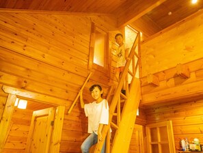 A wooden cabin interior with a central staircase, wooden walls, and a door on the left.
