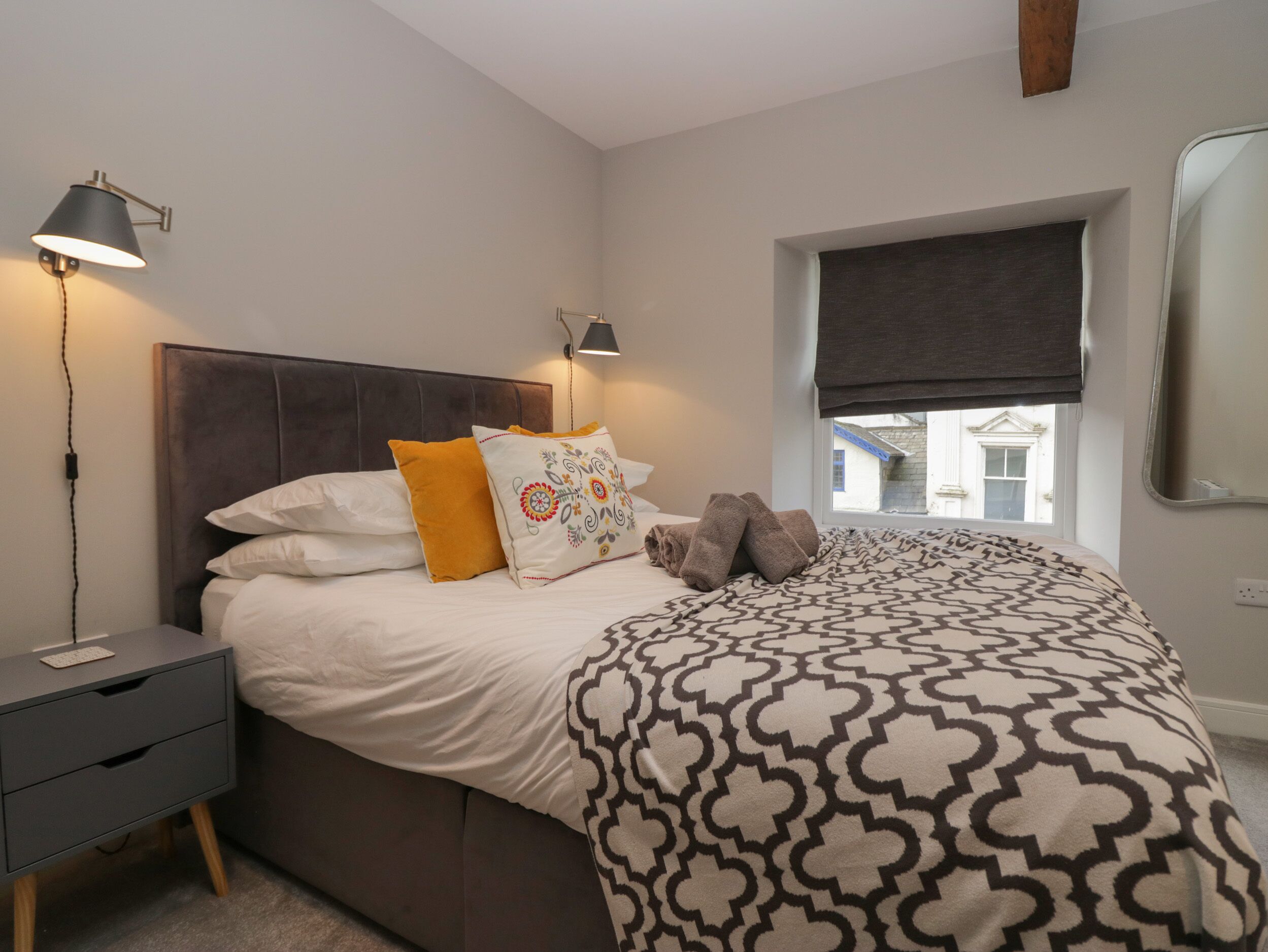A neatly made bed with patterned bedding, a bedside lamp, and a window with a view of buildings.