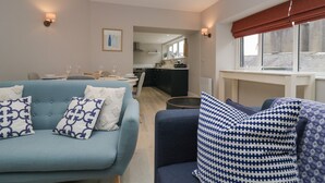 Un salon moderne avec un canapé et un fauteuil, un coin repas avec une table dressée pour deux personnes, et une cuisine en arrière-plan.