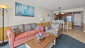 Ein Wohnzimmer mit einem rosa Sofa, gemusterten Sesseln, einem Holztisch und einem Gemälde mit Meeresblick.