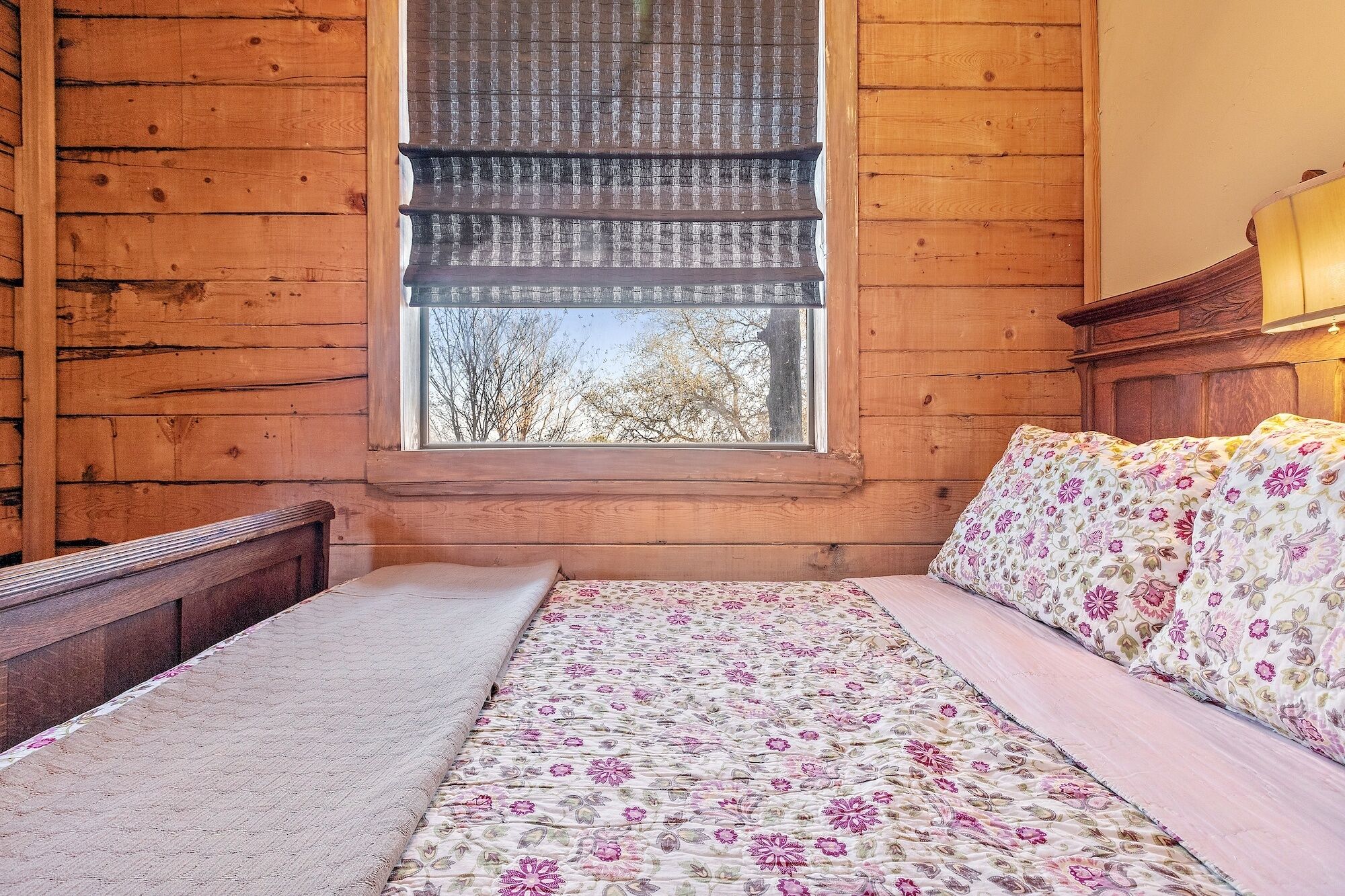 A wooden cabin room with a bed, a window with a view of trees, and a wooden headboard.