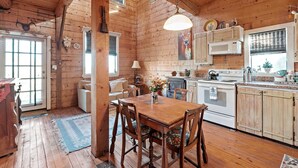 A rustic cabin interior with a wooden dining table, chairs, a kitchen with a stove and microwave, and a large window. - Mighty Oak Log Cabin 2 Bedroom Cabin by RedAwning (Fredericksburg)