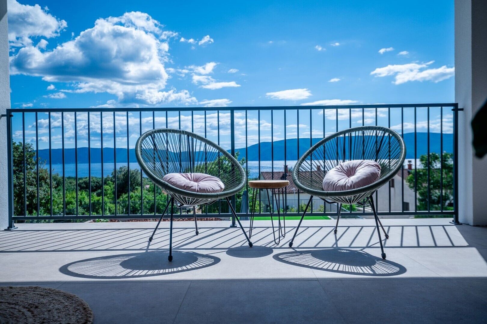 A balcony with two wicker chairs and a small table, overlooking a scenic view of mountains and a body of water.