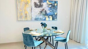 A modern dining area with a glass table and blue chairs, a large abstract painting on the wall, and a view of the outdoors through sheer curtains. - Luxury Dubai Creek Harbour Waterfront Apartment (Dubai)