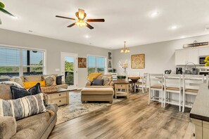 A spacious living room with a sofa, ottoman, and dining area with chairs and a table. There is a ceiling fan, a kitchen area with cabinets, and a window with blinds. - Exquisite Pet Friendly Condo Conveniently Located Close to the Fun In Branson (Branson West)