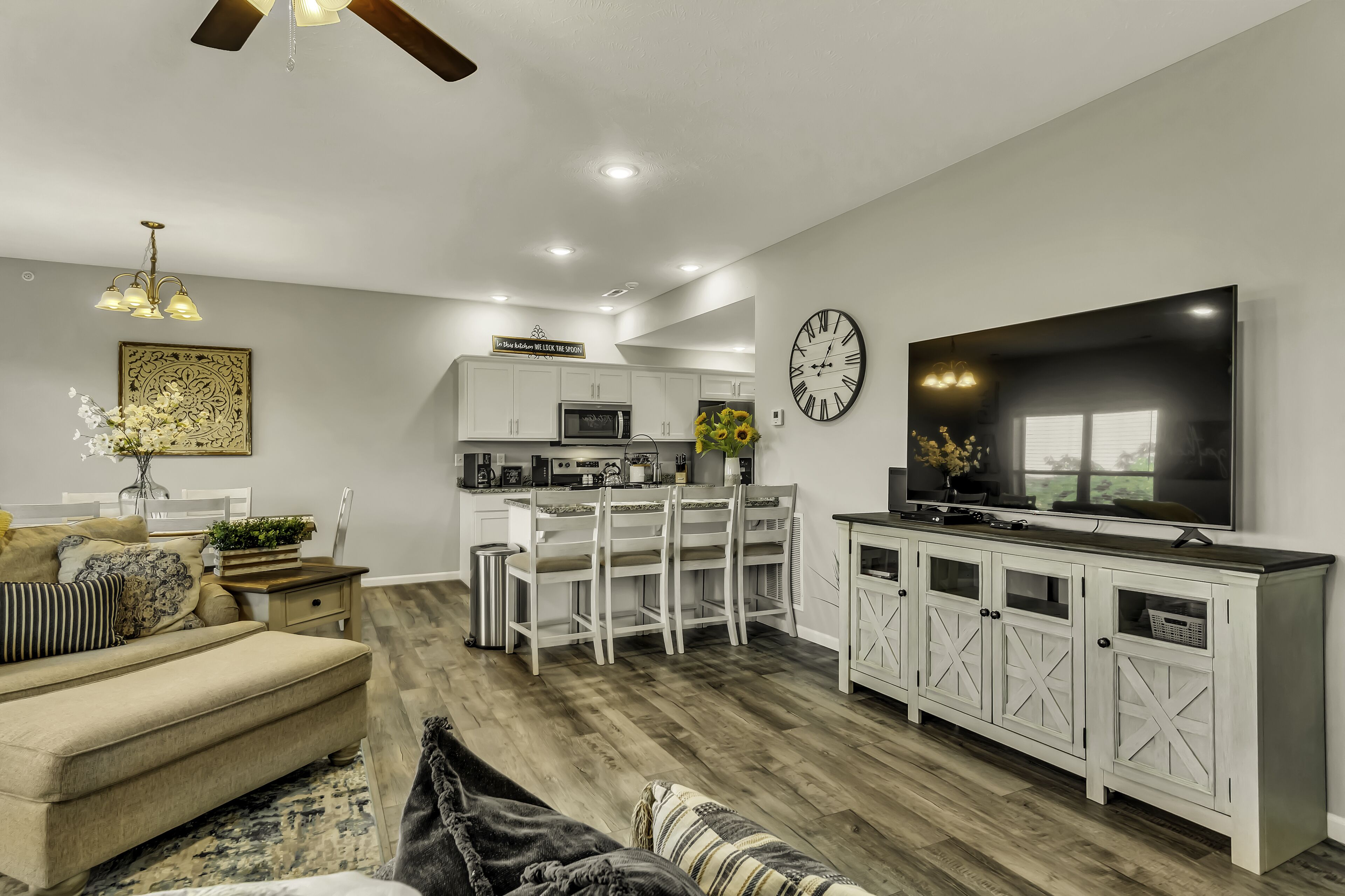 A modern living room with a sofa, a coffee table, and a TV. There is a kitchen area with white cabinets and a dining table.