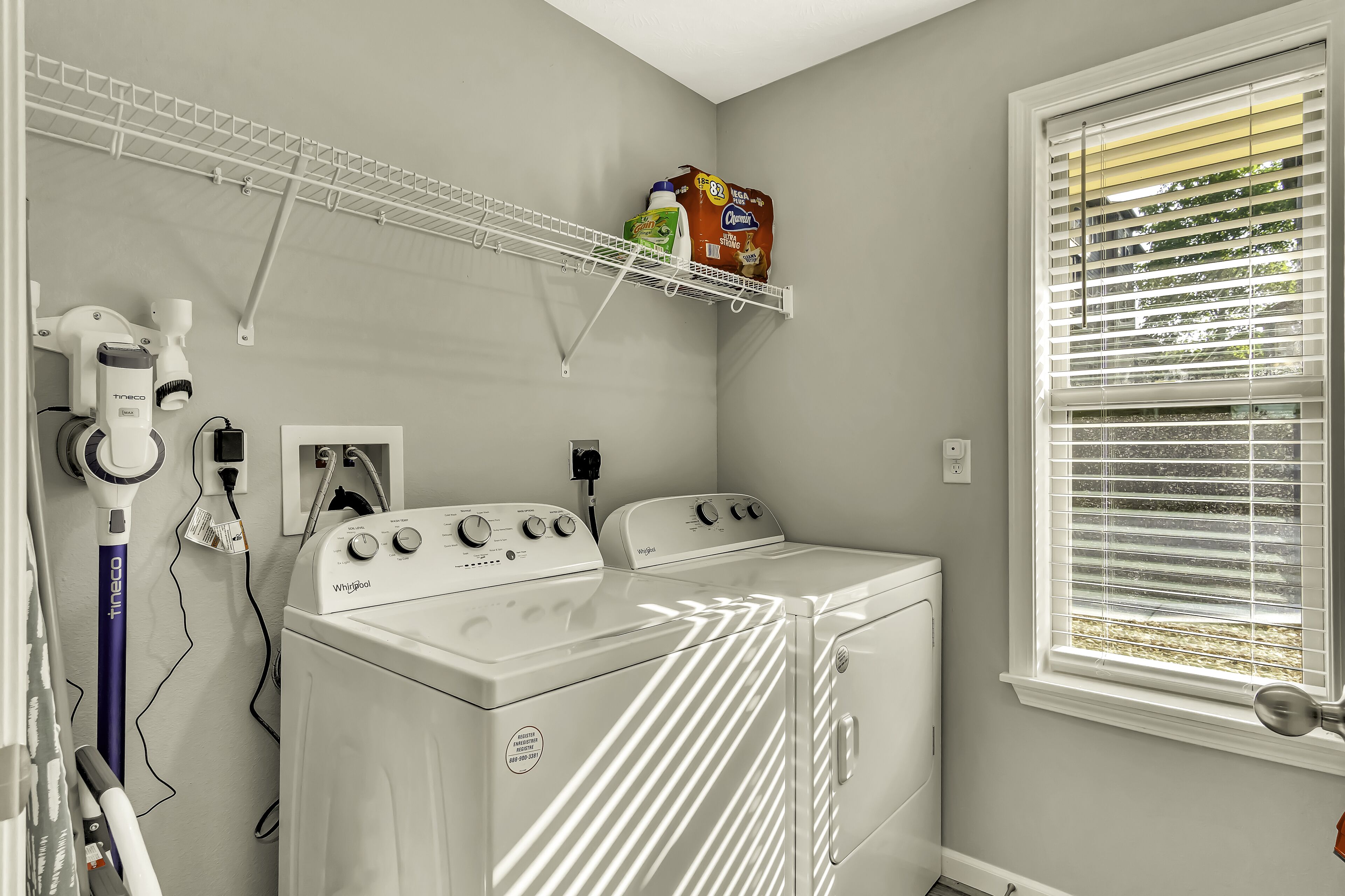 Une buanderie équipée d’un lave-linge et d’un sèche-linge à chargement frontal, d’un étendoir fixé au plafond et d’une fenêtre avec des stores.