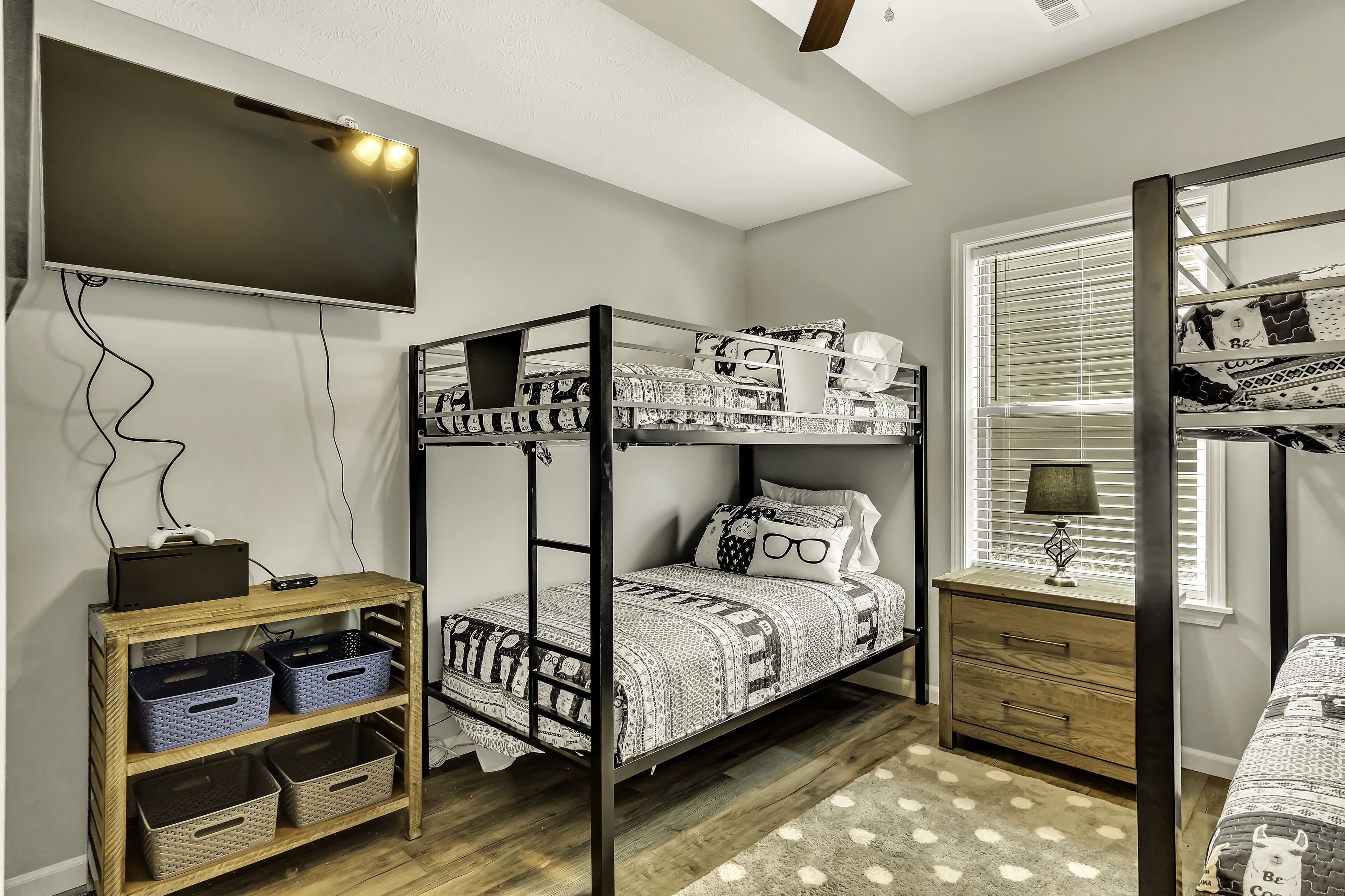 A bunk bed room with a TV, wooden storage unit, and a window with blinds.
