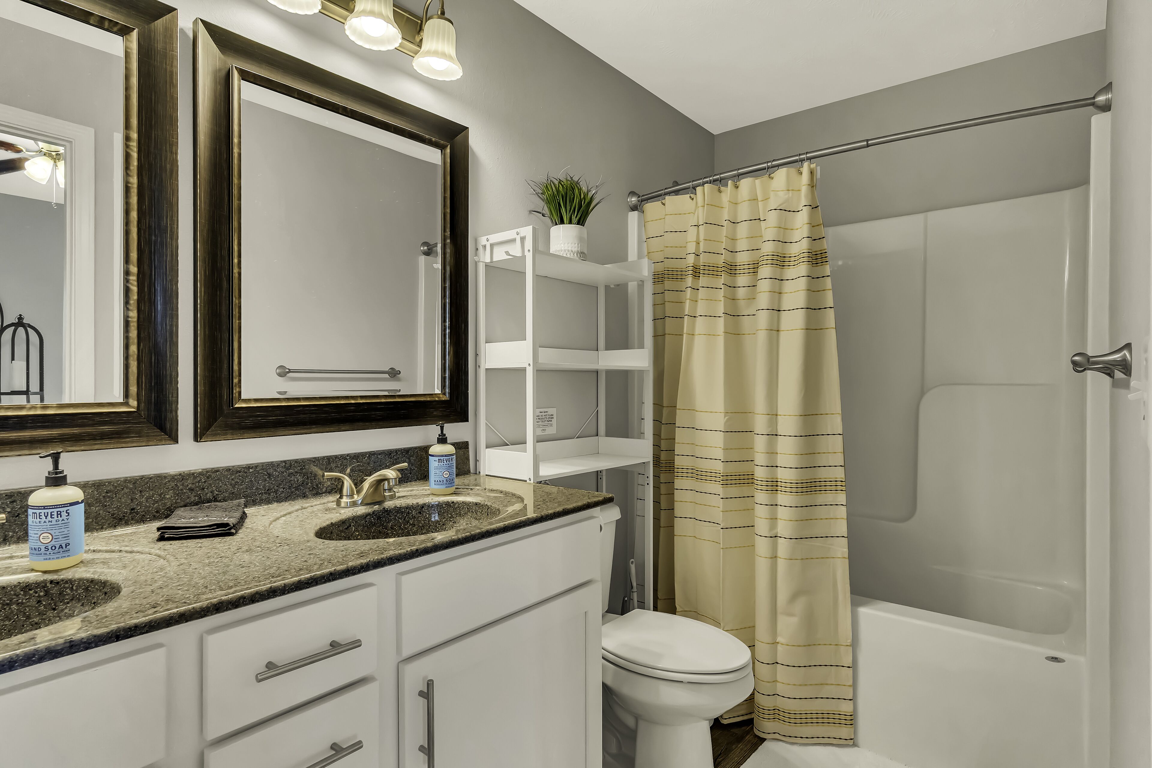 A bathroom with a double sink vanity, a toilet, a shower with a striped curtain, and a mirror with a wooden frame.
